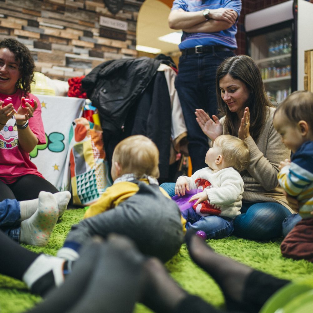 Mothers and their baby children are in a sensory play group. They are singing and dancing together in a circle on the floor.